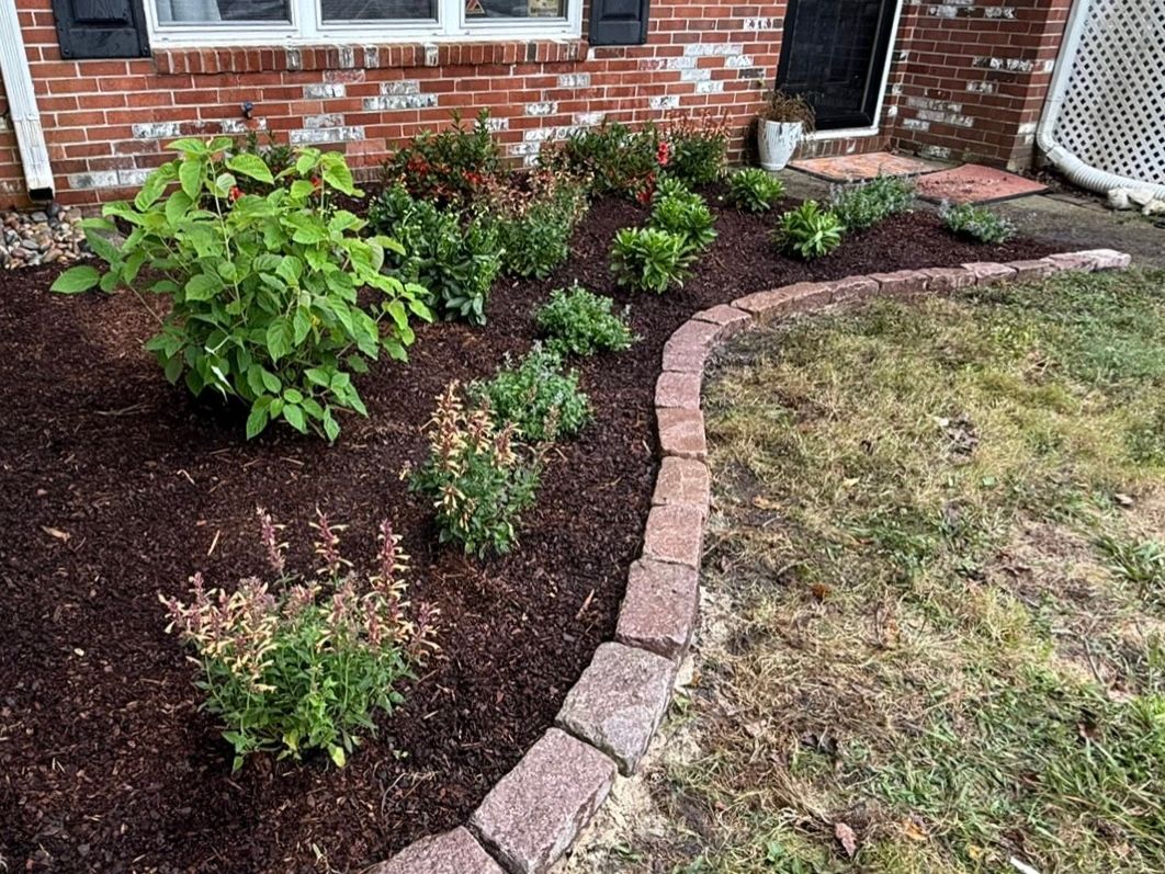 Mulched garden bed with shrubs and flowers beside a brick house, edged by a curved brick border