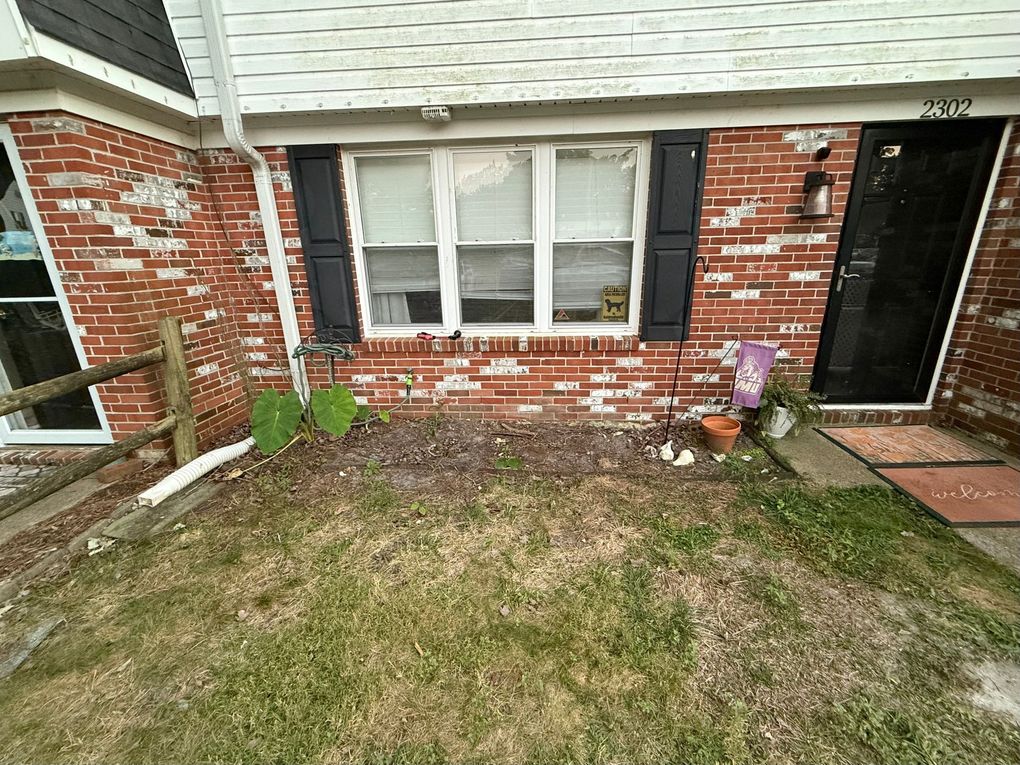 Front yard of a brick house with a small lawn, white window, and a black front door with potted flowers.