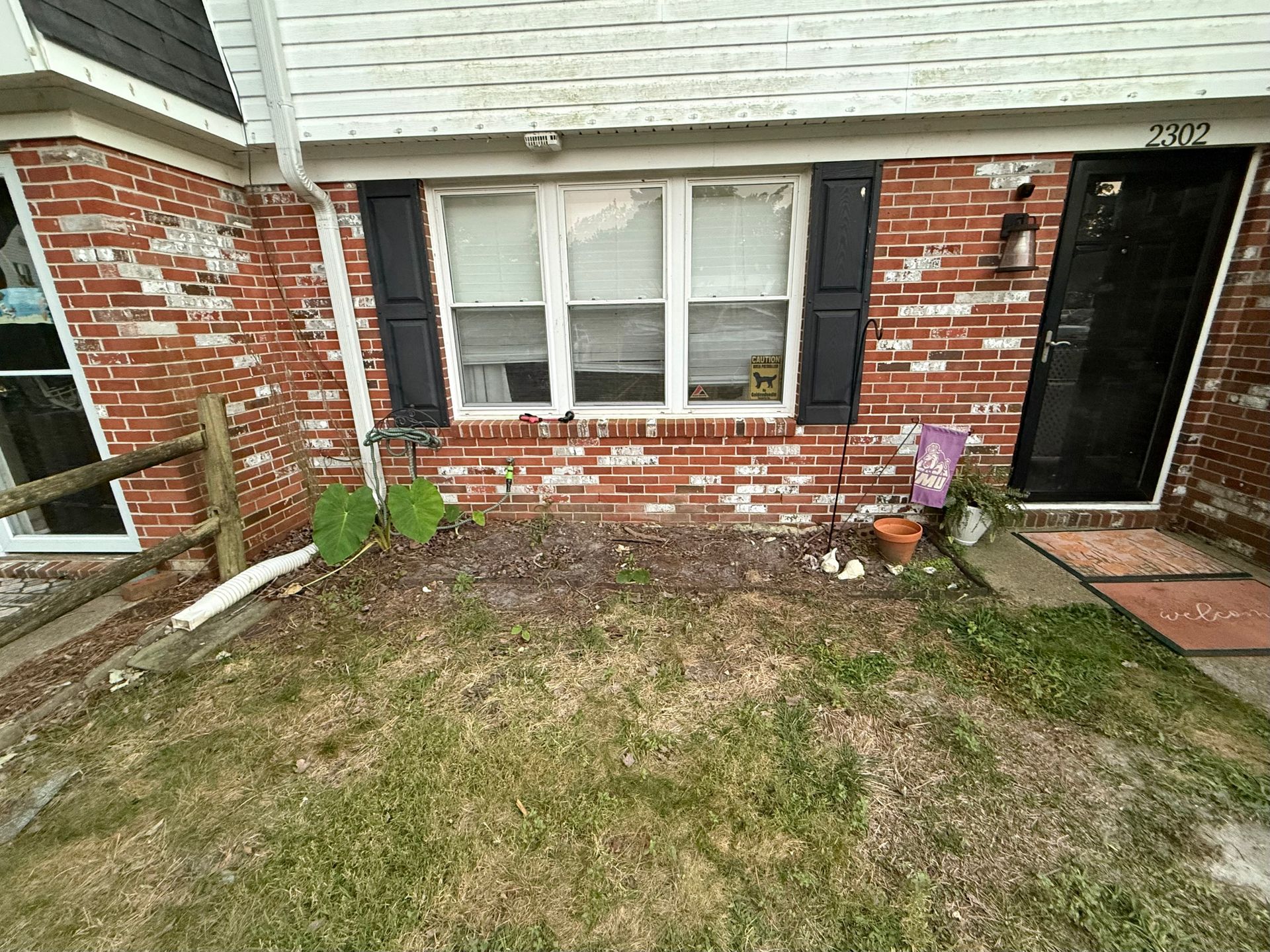Brick house front with a window, black door, small porch, and sparse front lawn with potted plants.