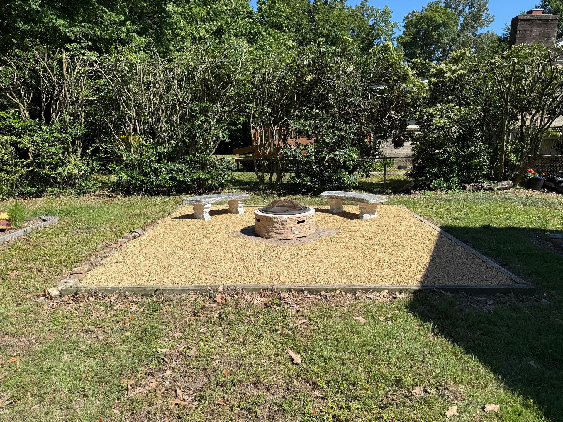 Backyard fire pit area with gravel seating pad and stone rings, surrounded by grass and trees
