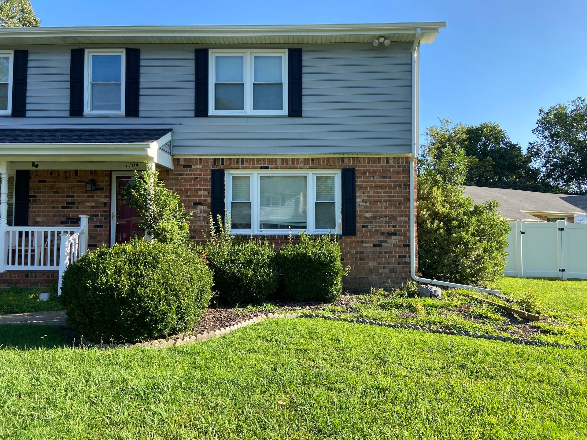 Suburban two-story brick and siding house with front porch, shrubs, green lawn, and detached garage.