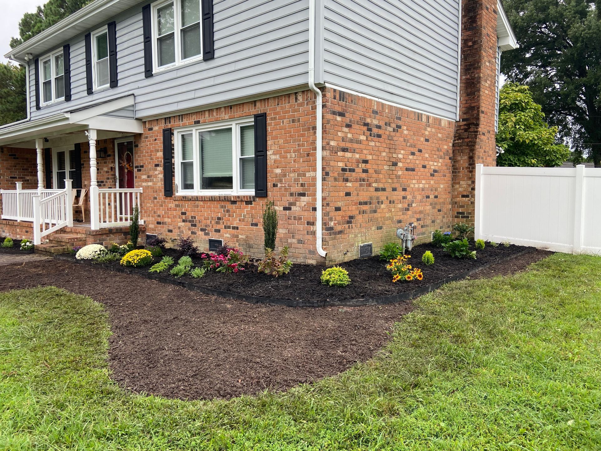 Two-story brick and gray house with a white fence and fresh mulch landscaping in front