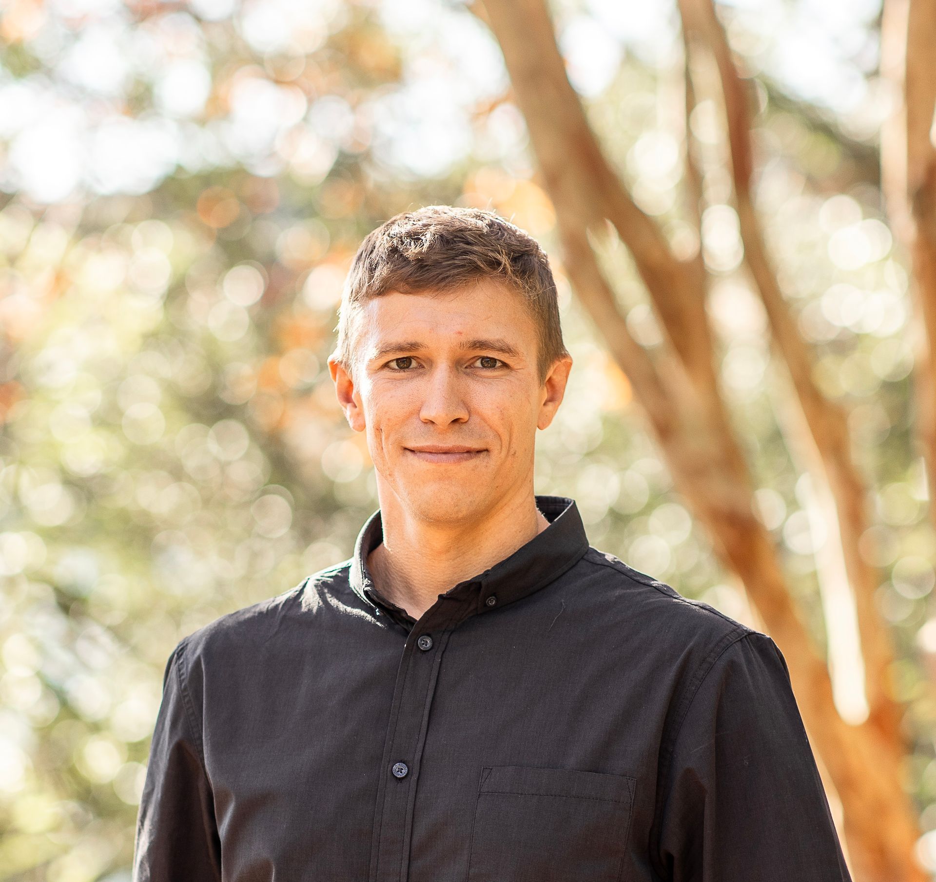 Portrait of a smiling man in a black shirt standing outdoors with blurred trees in the background