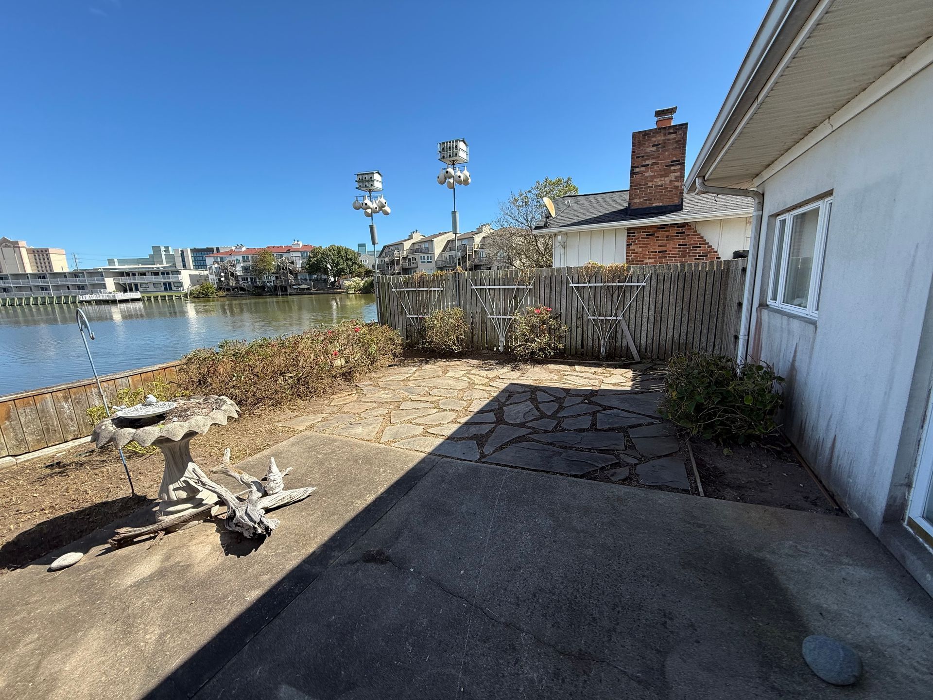 Backyard patio beside a waterfront, with a fence, white house wall, and blue sky.