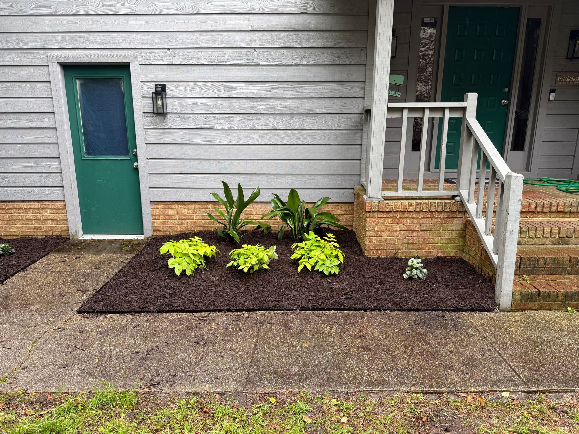 Front of a house with a green door, porch railing, and a small planted garden bed by the sidewalk