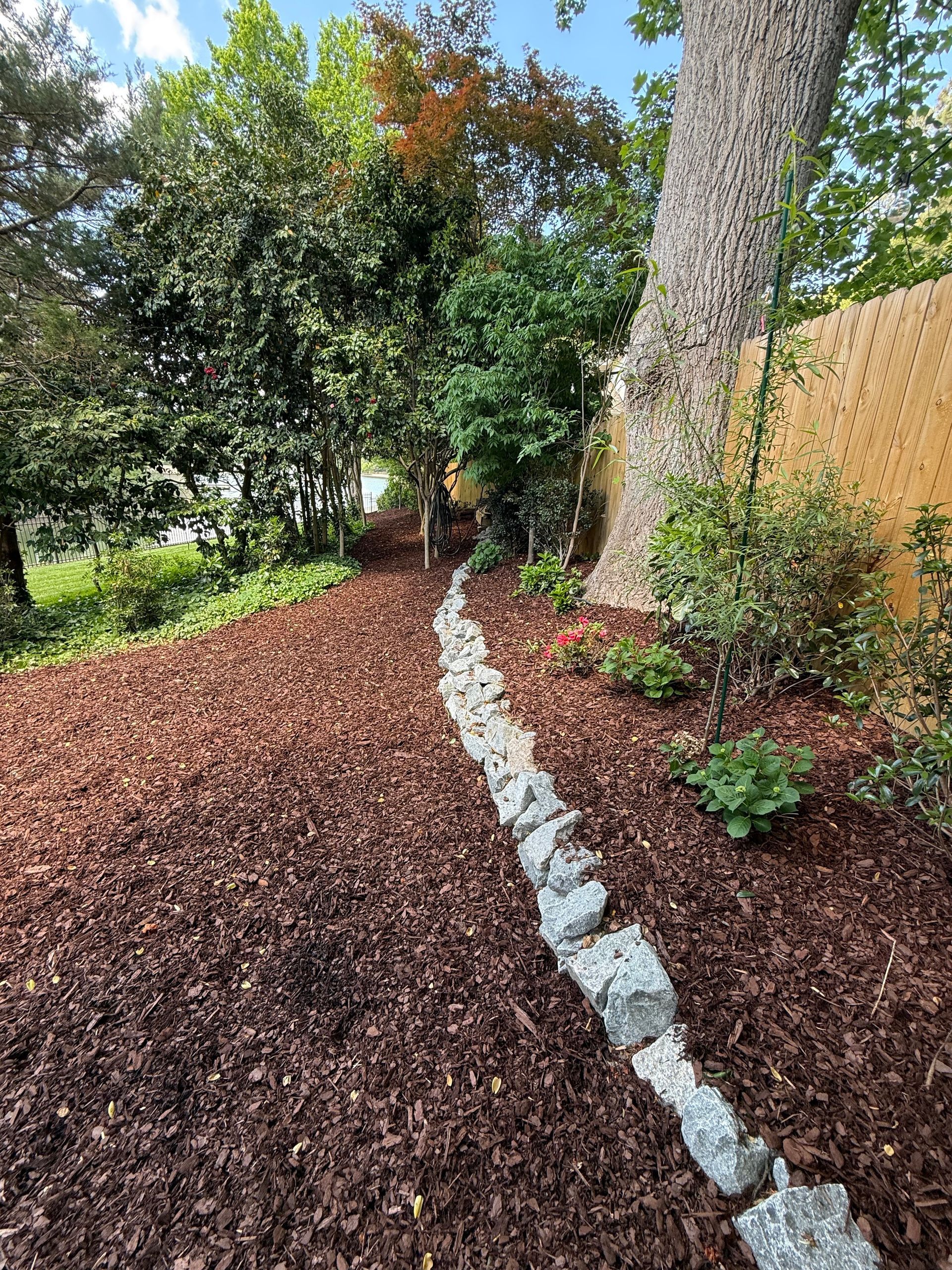 Mulched garden bed with stone border beside a tree and wooden fence in a backyard