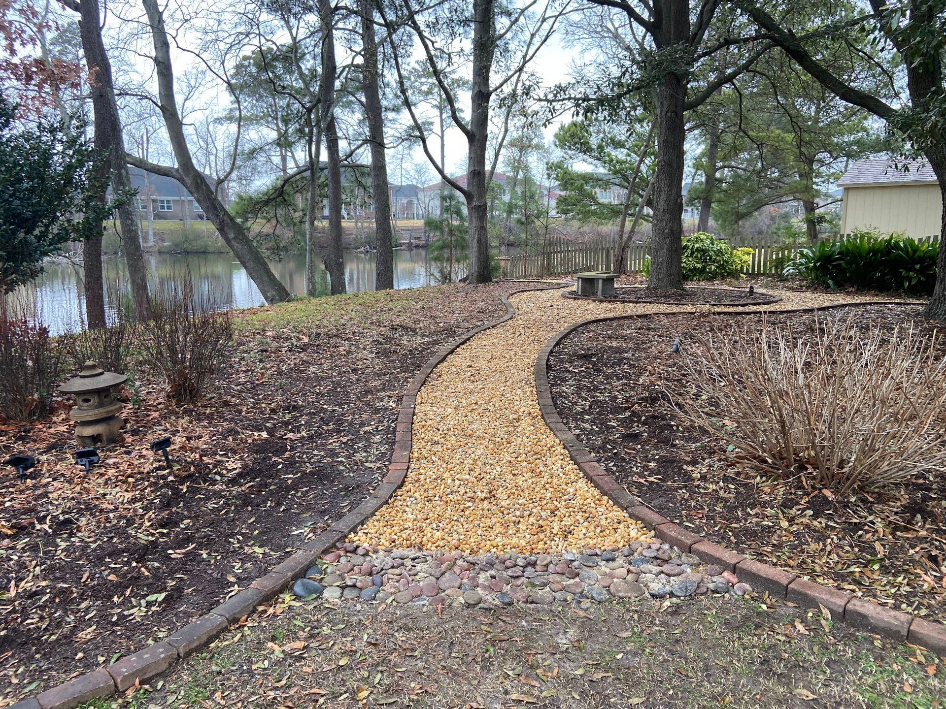 Curved gravel garden path through leafless trees beside a lake and house