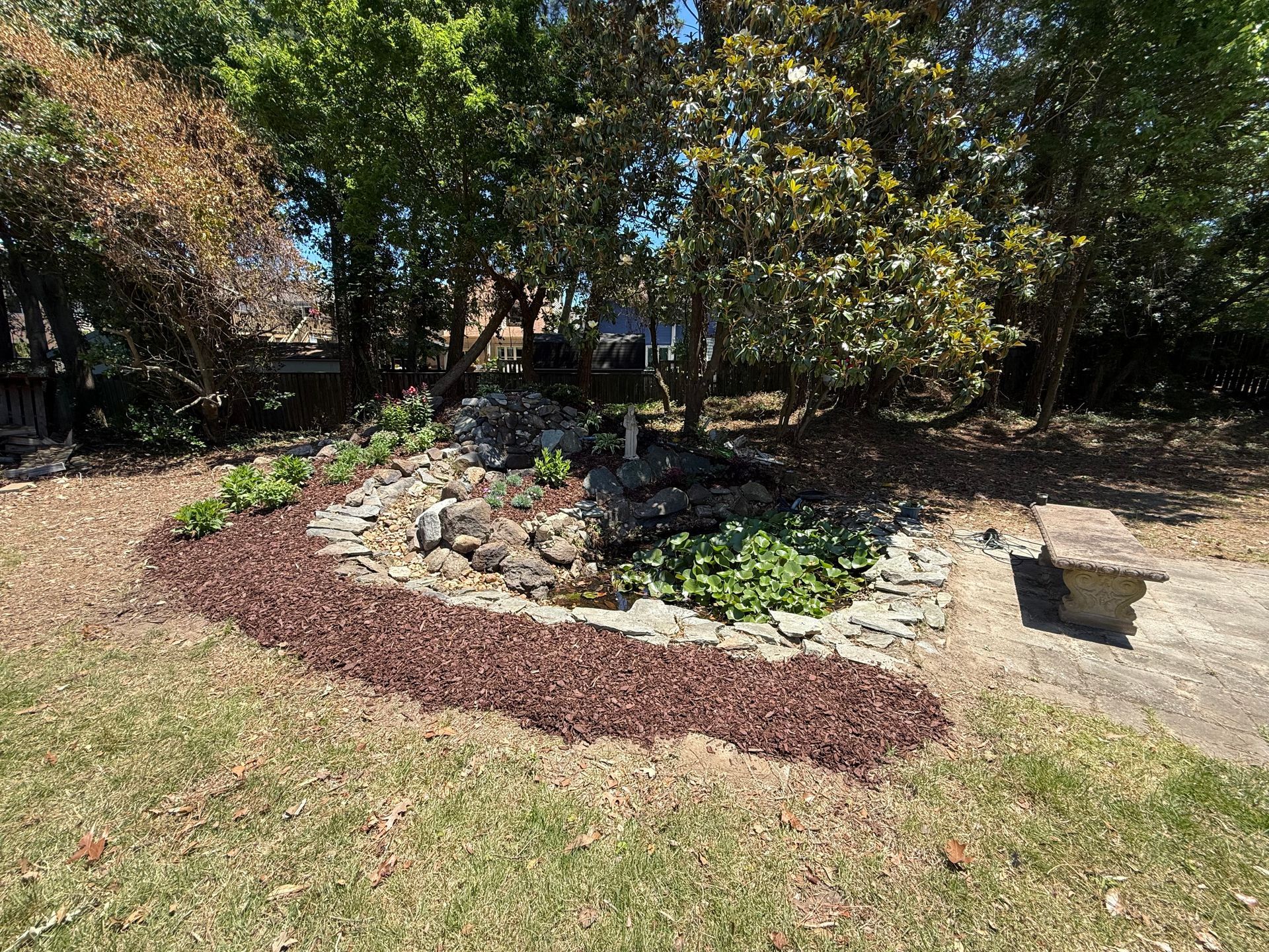 Backyard garden bed with rocks, mulch, and green plants beside a lawn and trees