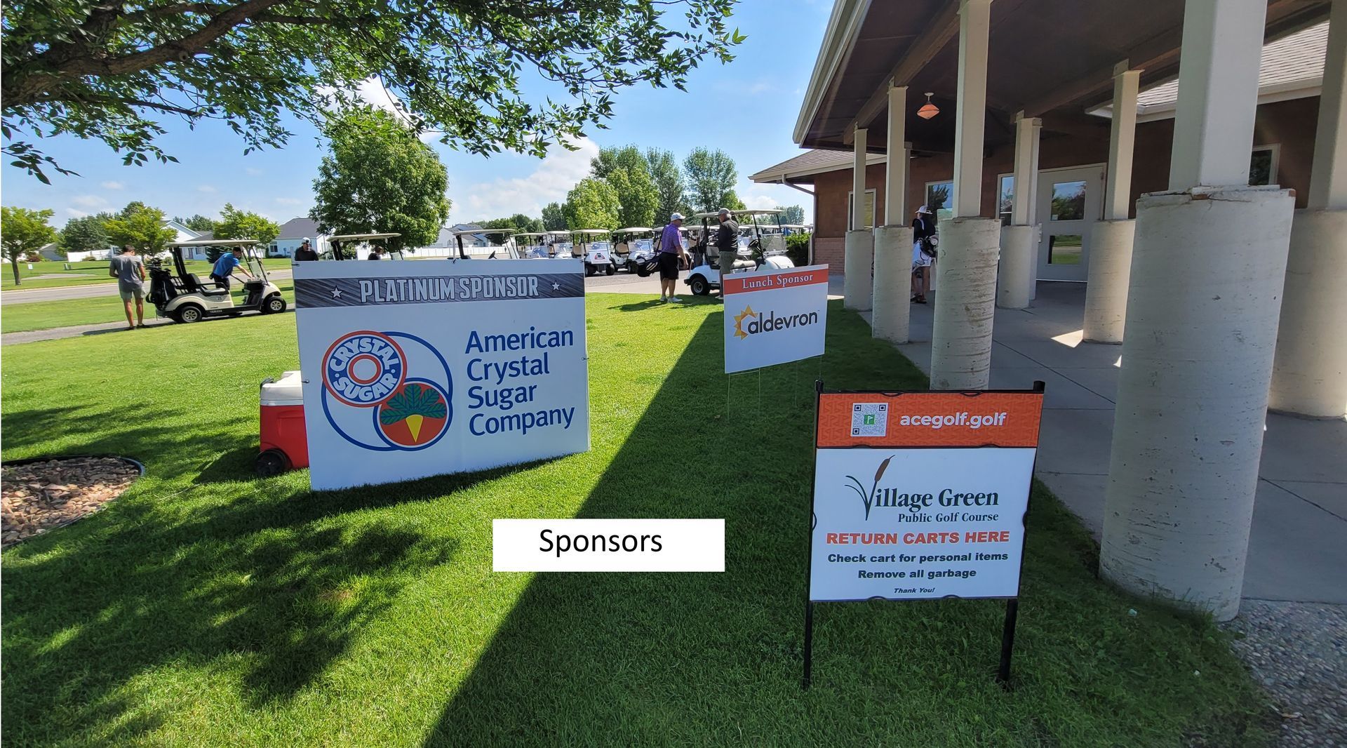 Event sponsors' signs on a grassy lawn with golf carts in the background. A building is to the right.
