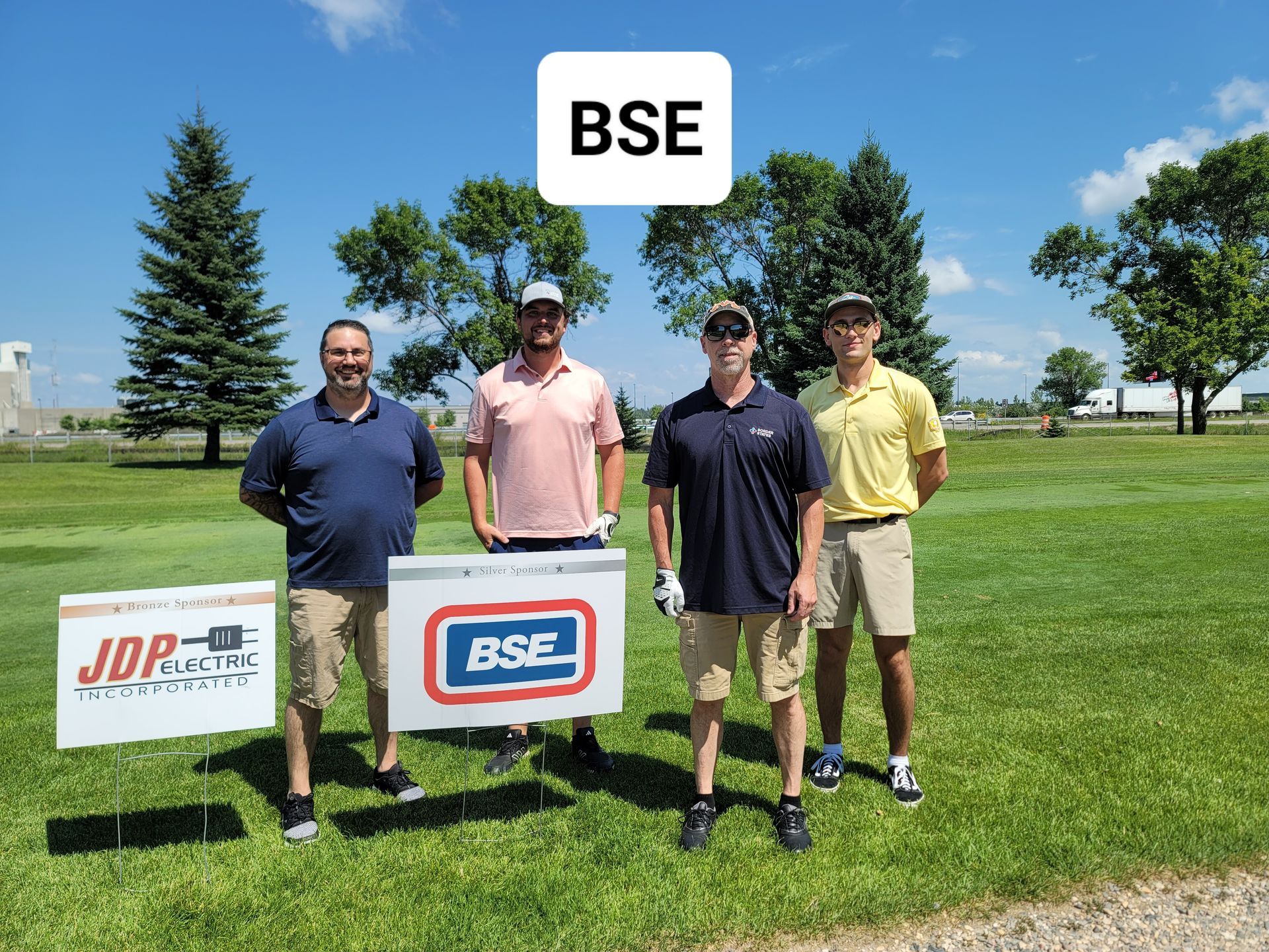 Four men pose with a sign on a golf course under a blue sky; sign says