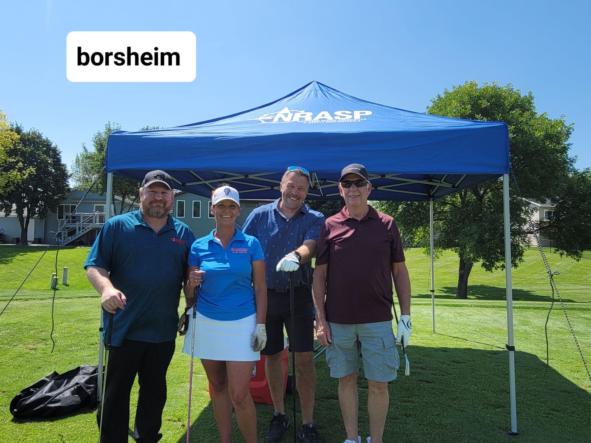 Four people under a blue tent on a golf course. They are holding golf clubs and smiling in the sun.