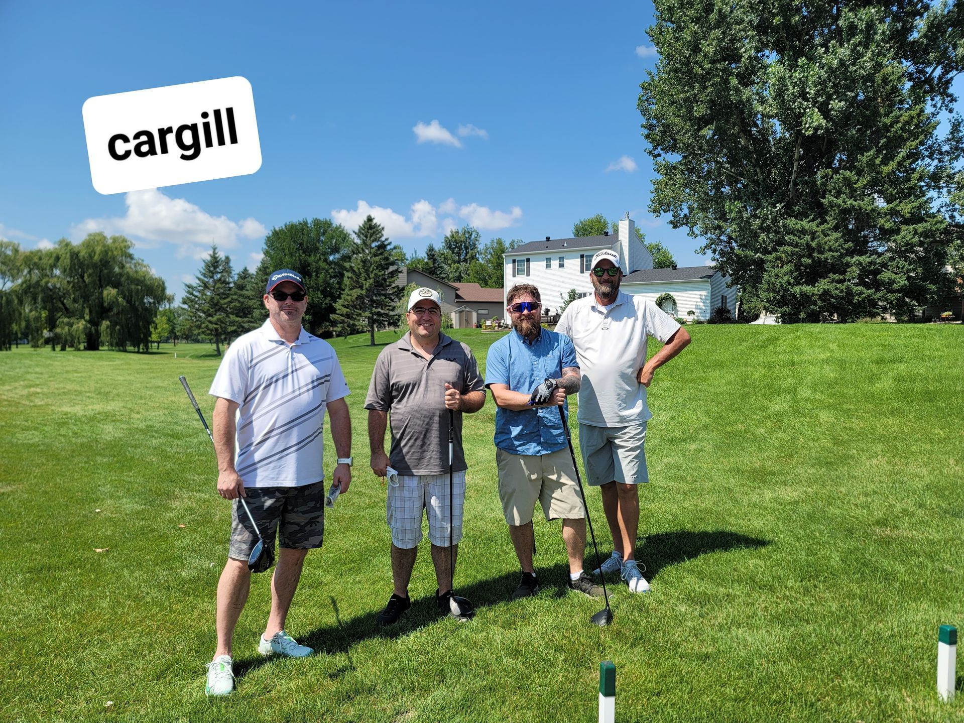 Four men on a golf course, holding clubs, under a blue sky,