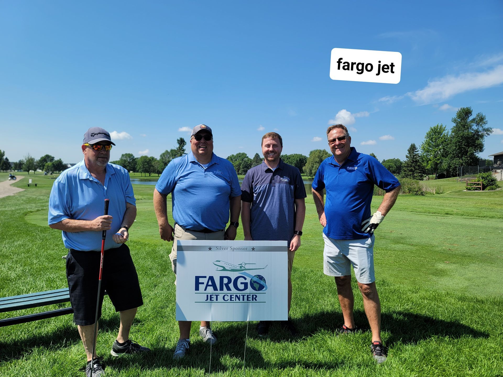 Four men pose on a golf course. They are standing behind a sign that reads