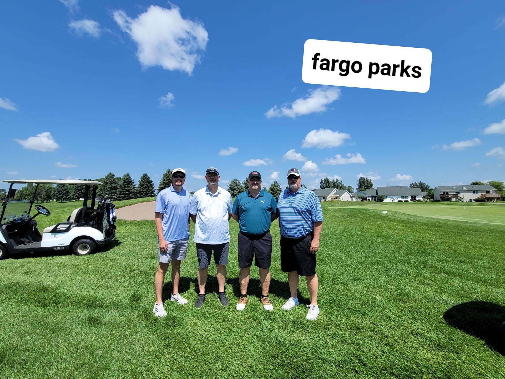 Four men pose on a golf course. A golf cart is parked nearby. Sunny day with blue sky.