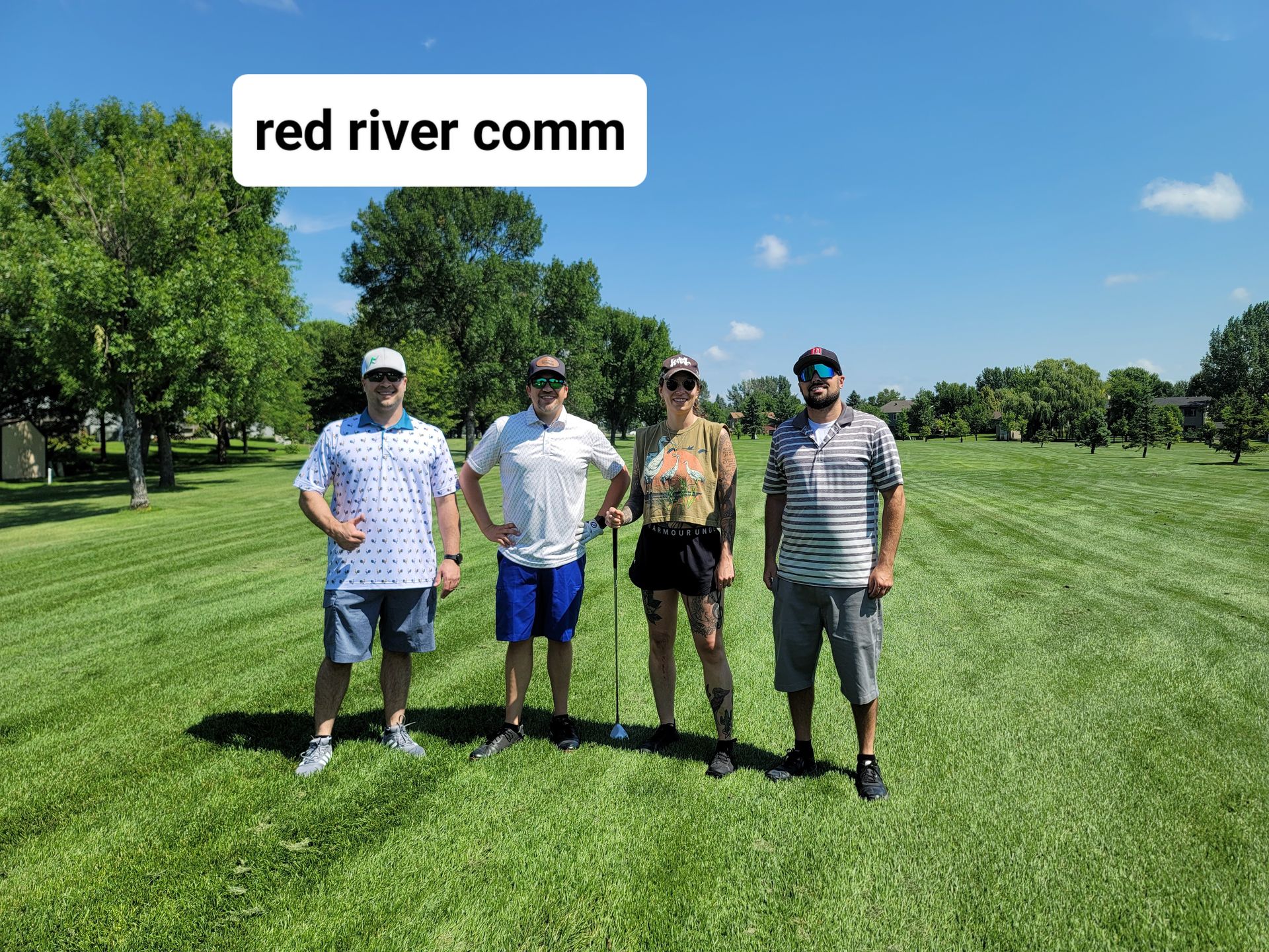 Four people standing on a green field under a blue sky, text reads