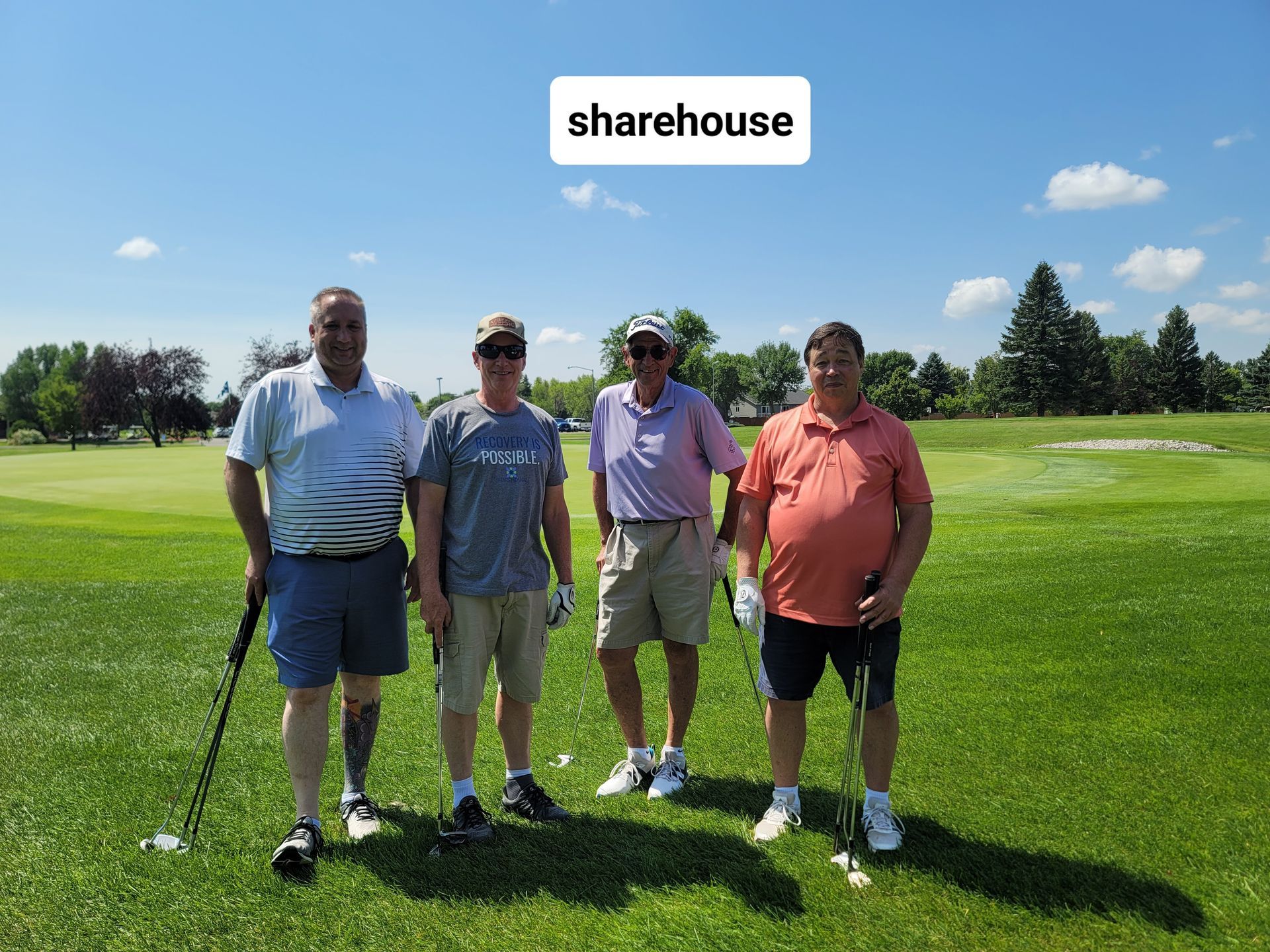 Four men on a golf course, standing on the grass with golf clubs, under a blue sky.