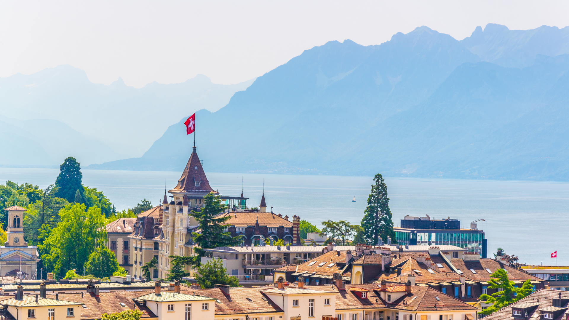 Swiss castle with red flag atop a tower, overlooking a lake and mountains.