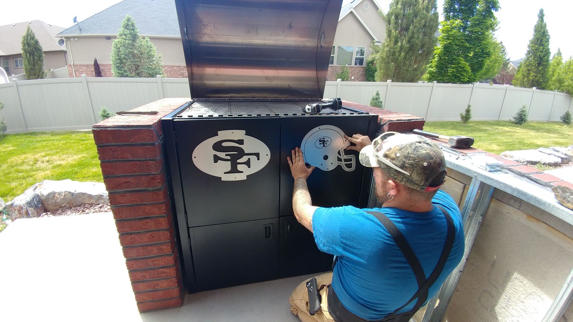 Man attaching a decoration to a black grill, built into a brick structure outdoors.