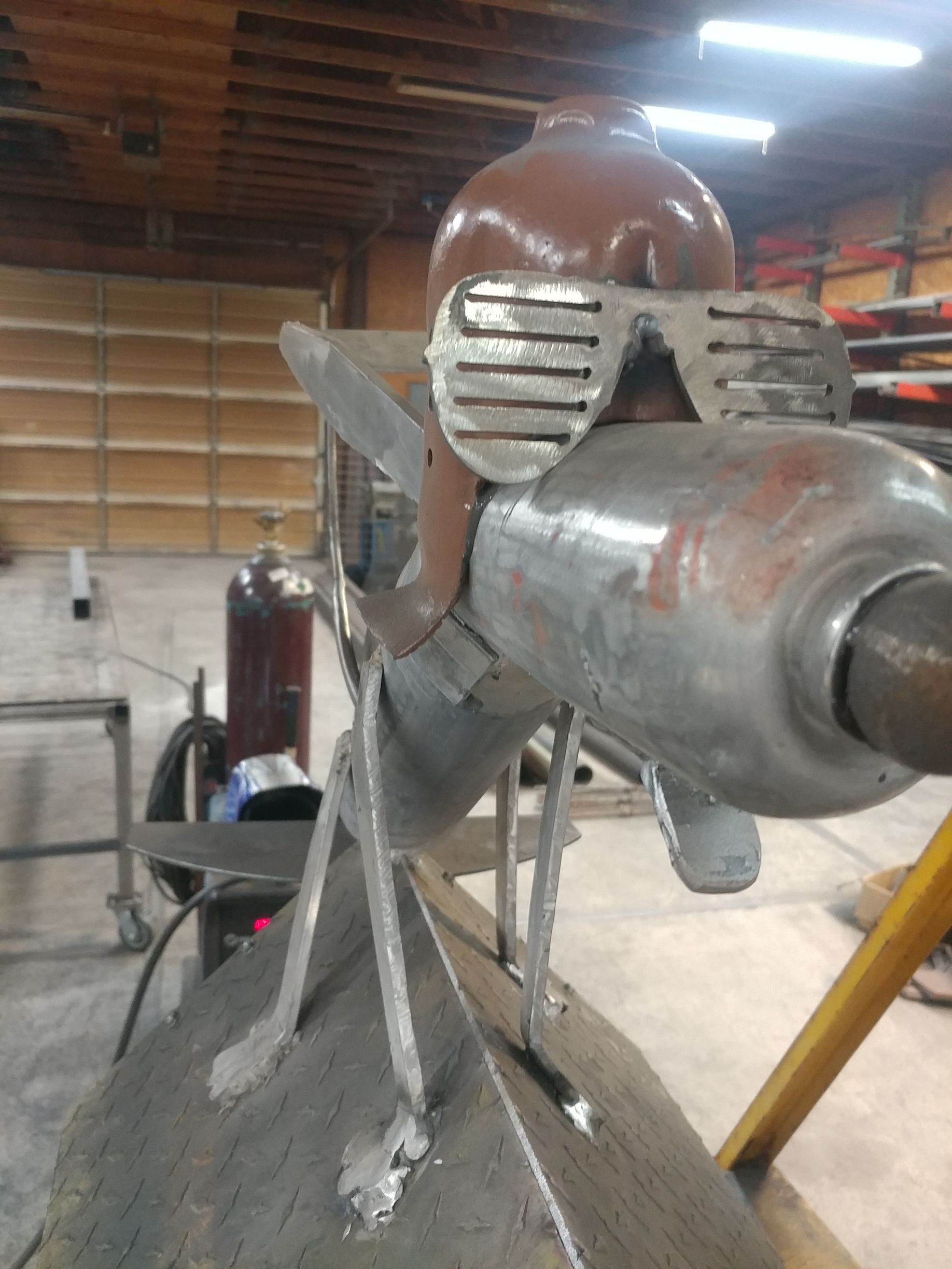 Welded metal sculpture of a creature with sunglasses, brown head, and tube-like body parts; inside a workshop.