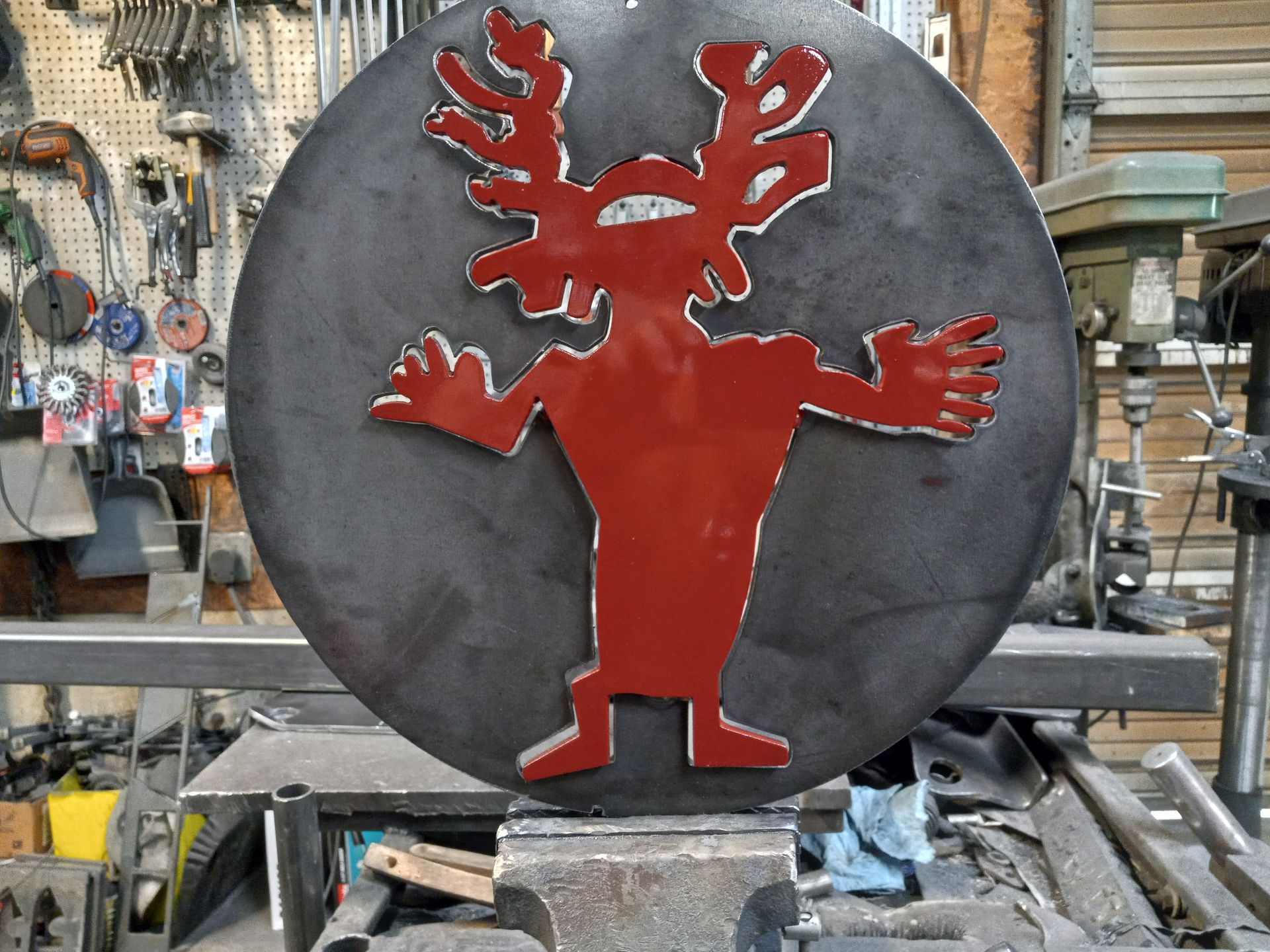 Red cartoon deer figure welded onto a dark metal circle, sitting on a metal workshop bench.