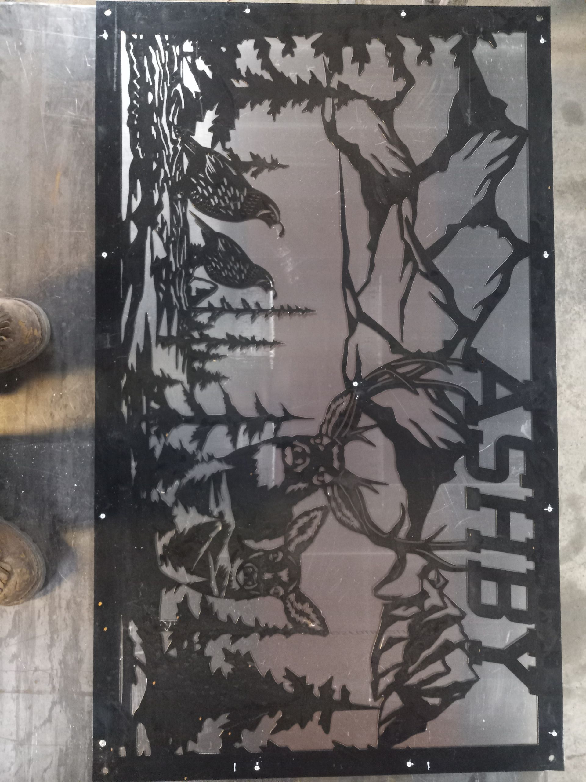 Laser-cut metal artwork featuring a deer, forest scene, two birds, and the name