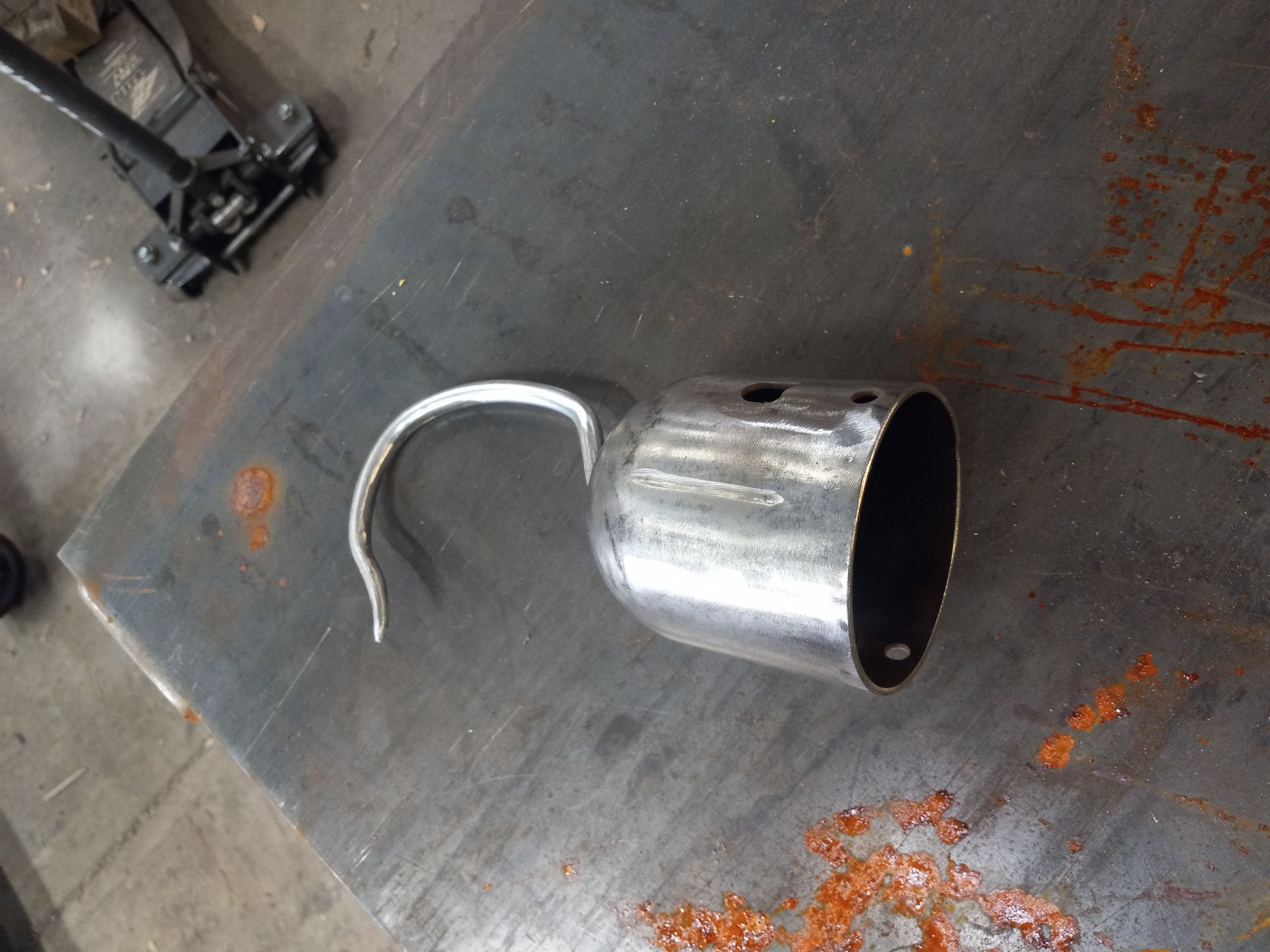 Metal hook-shaped object on a metal surface, possibly a pipe or part, with holes and a curved piece.