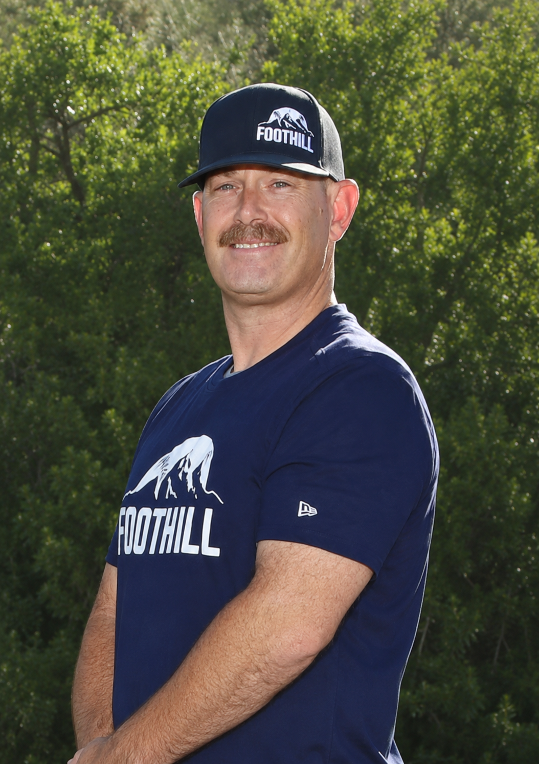 Man in green polo stands on a football field, arms crossed, stadium in background.
