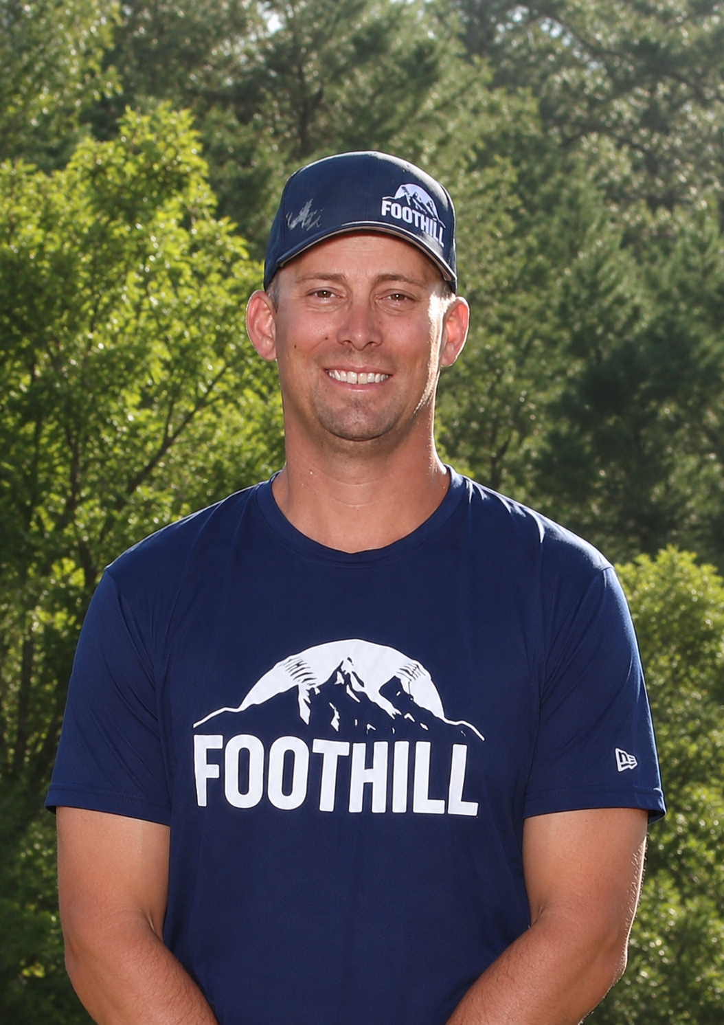 Man in green polo stands on a football field, arms crossed, stadium in background.