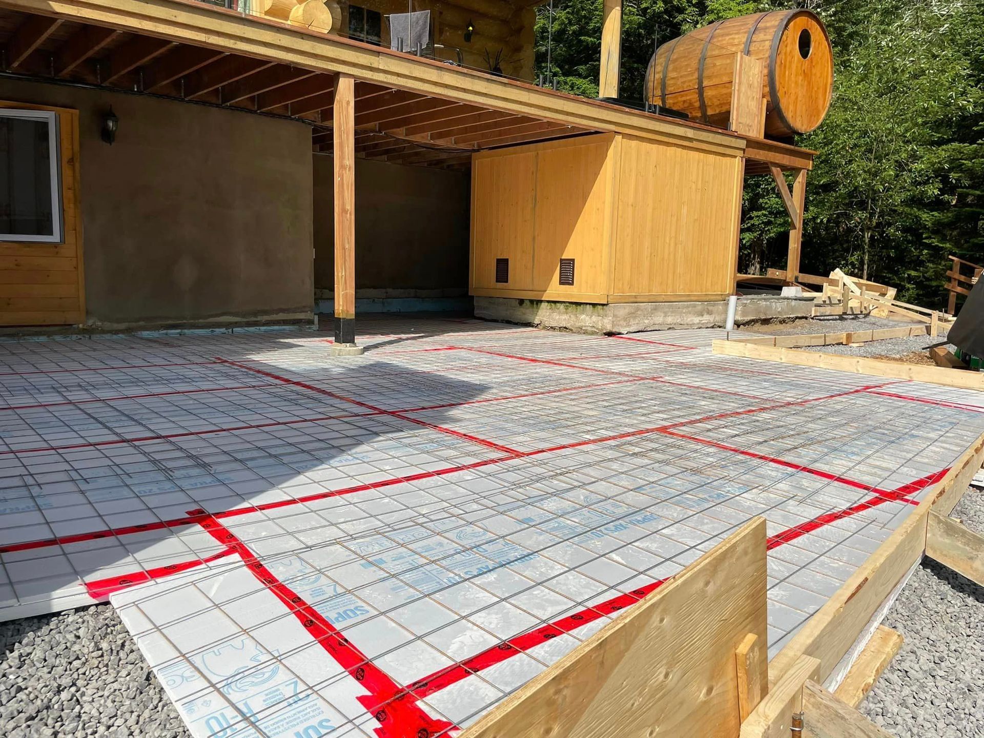 Terrasse extérieure en construction avec des tubes de chauffage radiants rouges posés sur l'isolant, structure en bois en arrière-plan.
