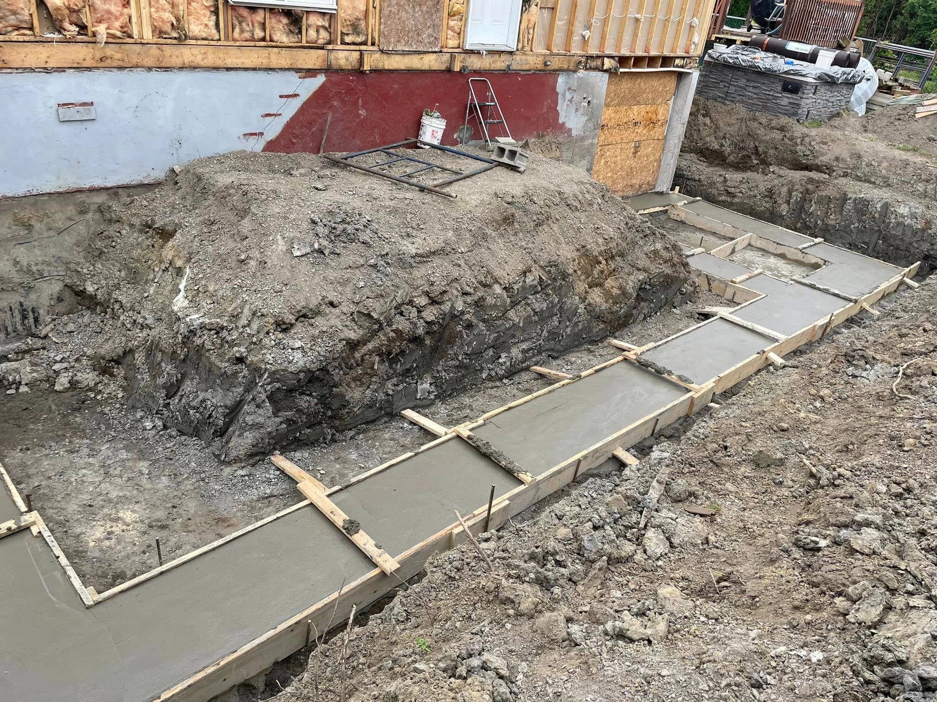 Chantier de construction avec semelles en béton coulé et coffrages en bois le long d'une tranchée ; terre et bâtiment en arrière-plan.