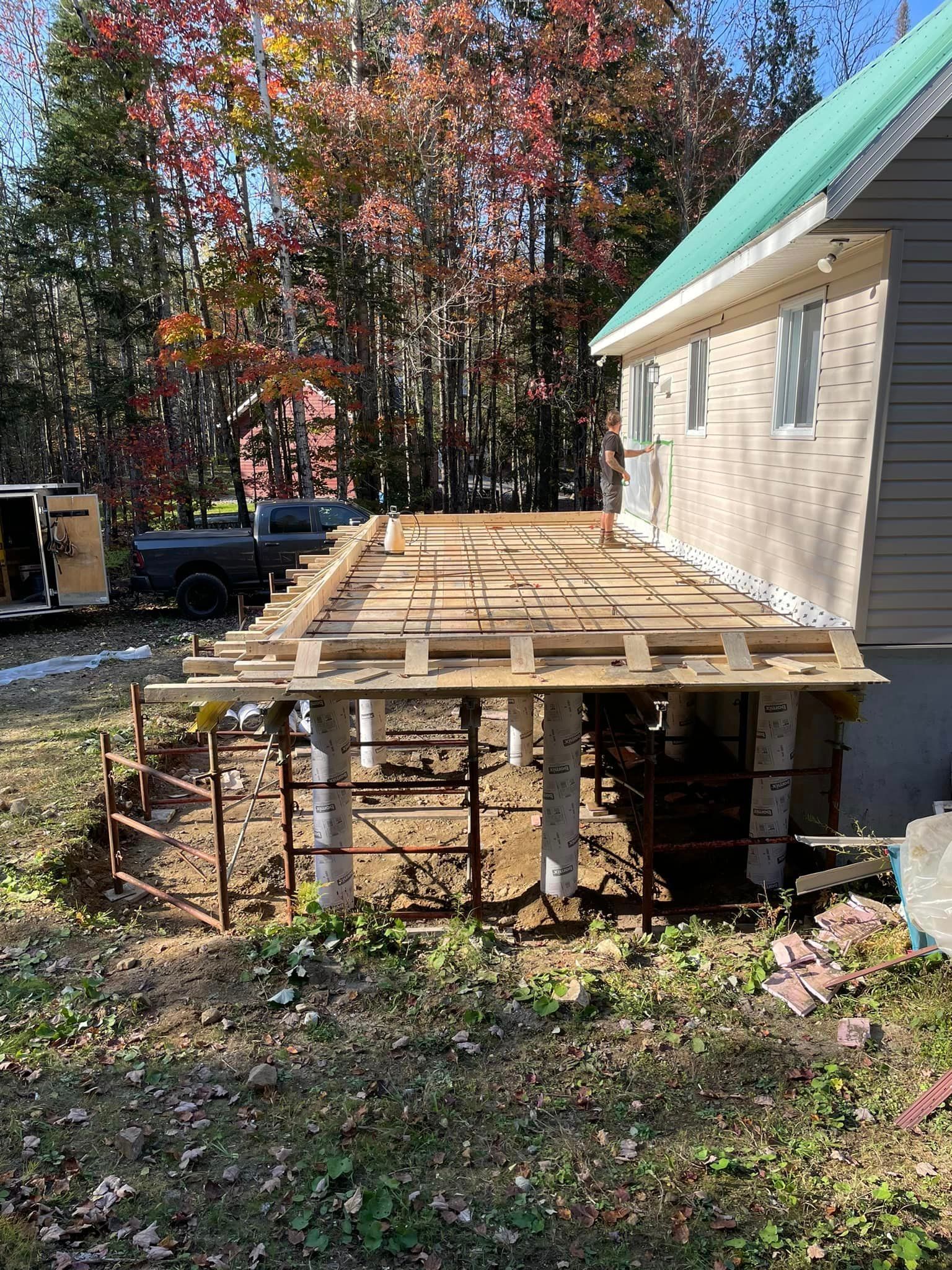 Construction d'une terrasse. Charpente en bois reposant sur des piliers en béton, à côté d'une maison au bardage gris.