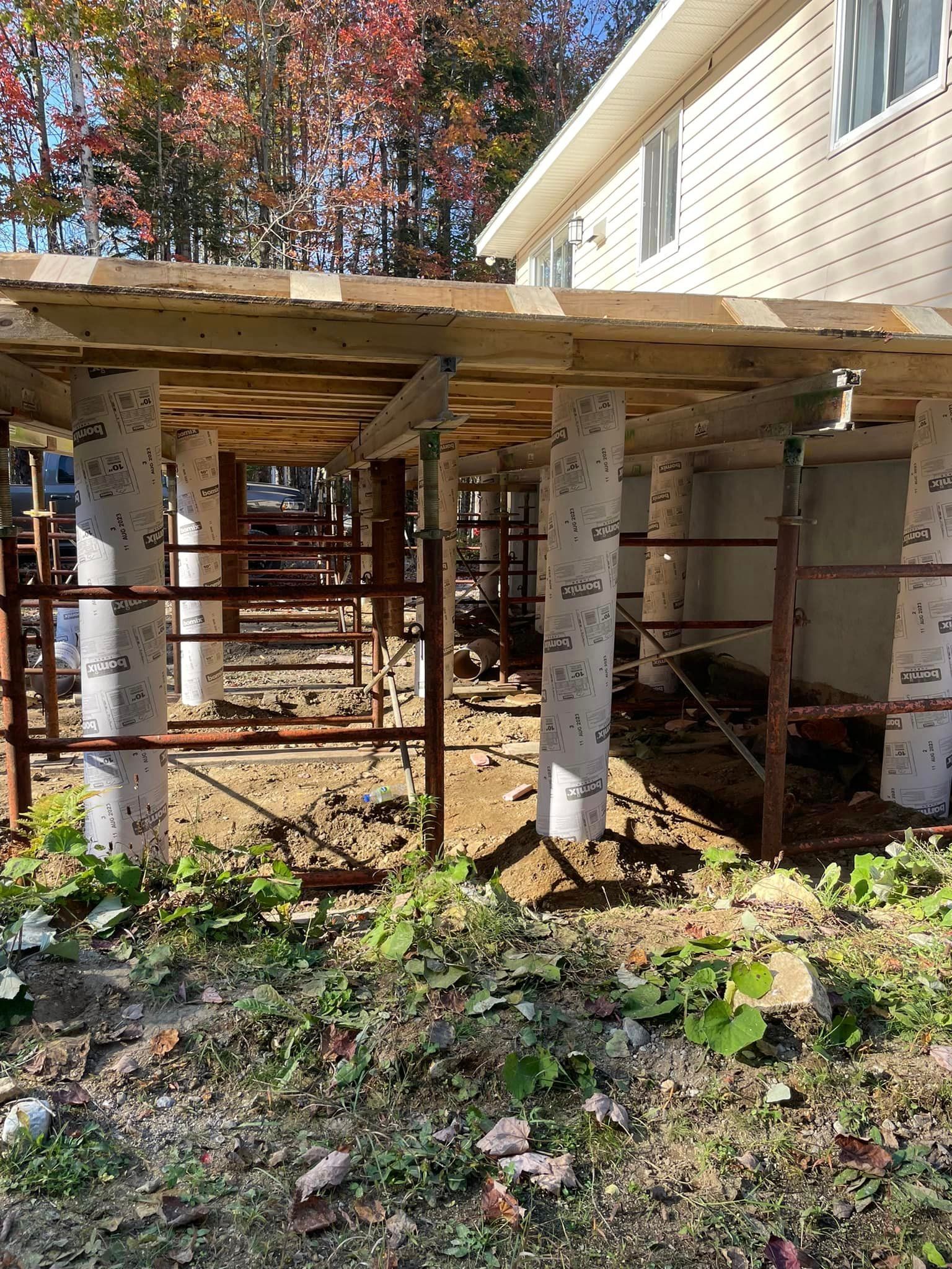 Construction d'une terrasse ; des colonnes en béton soutiennent une structure en bois à côté d'une maison.