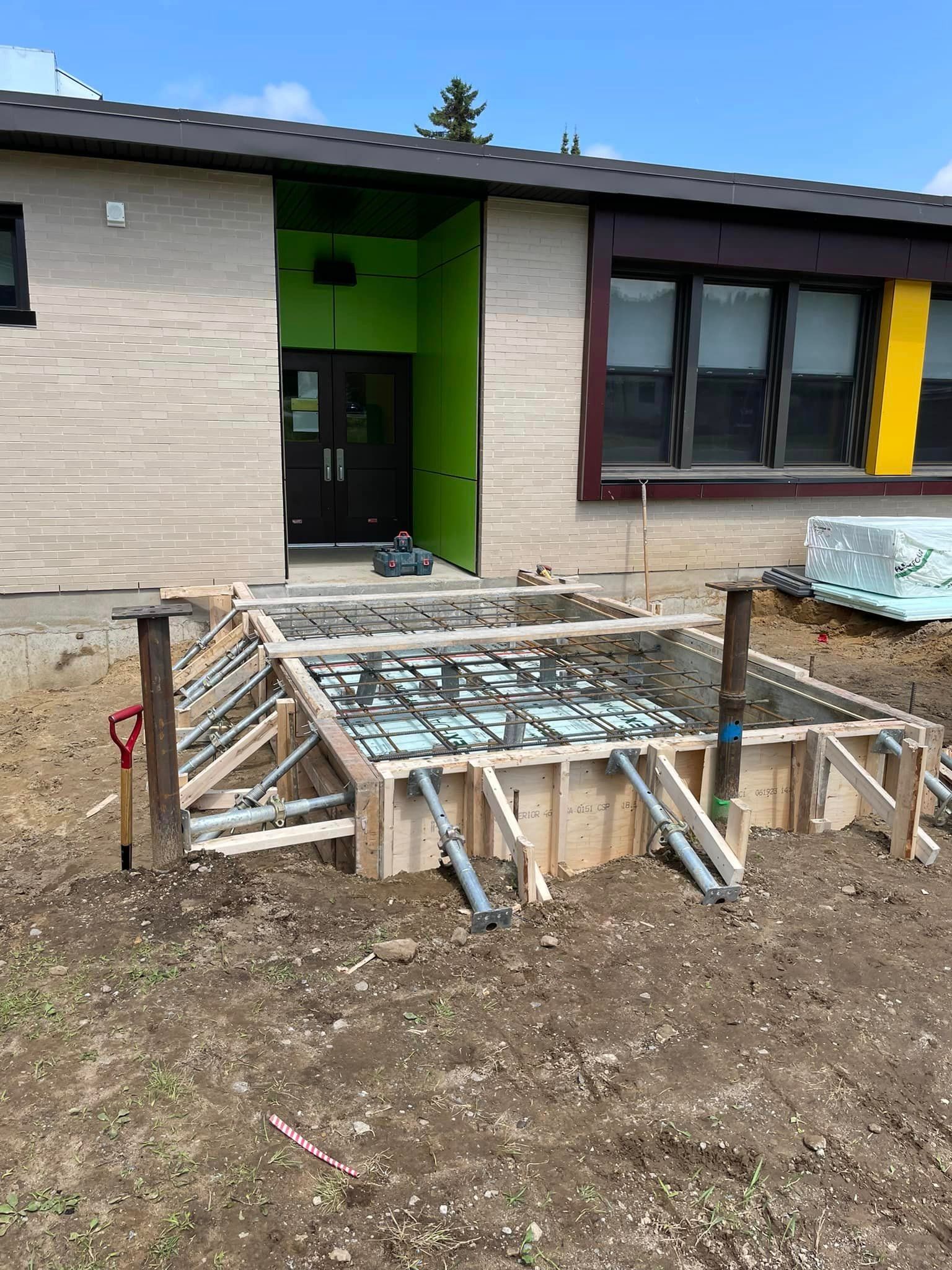 Chantier de construction devant un bâtiment. Un coffrage en bois avec des armatures métalliques est enfoui dans le sol.