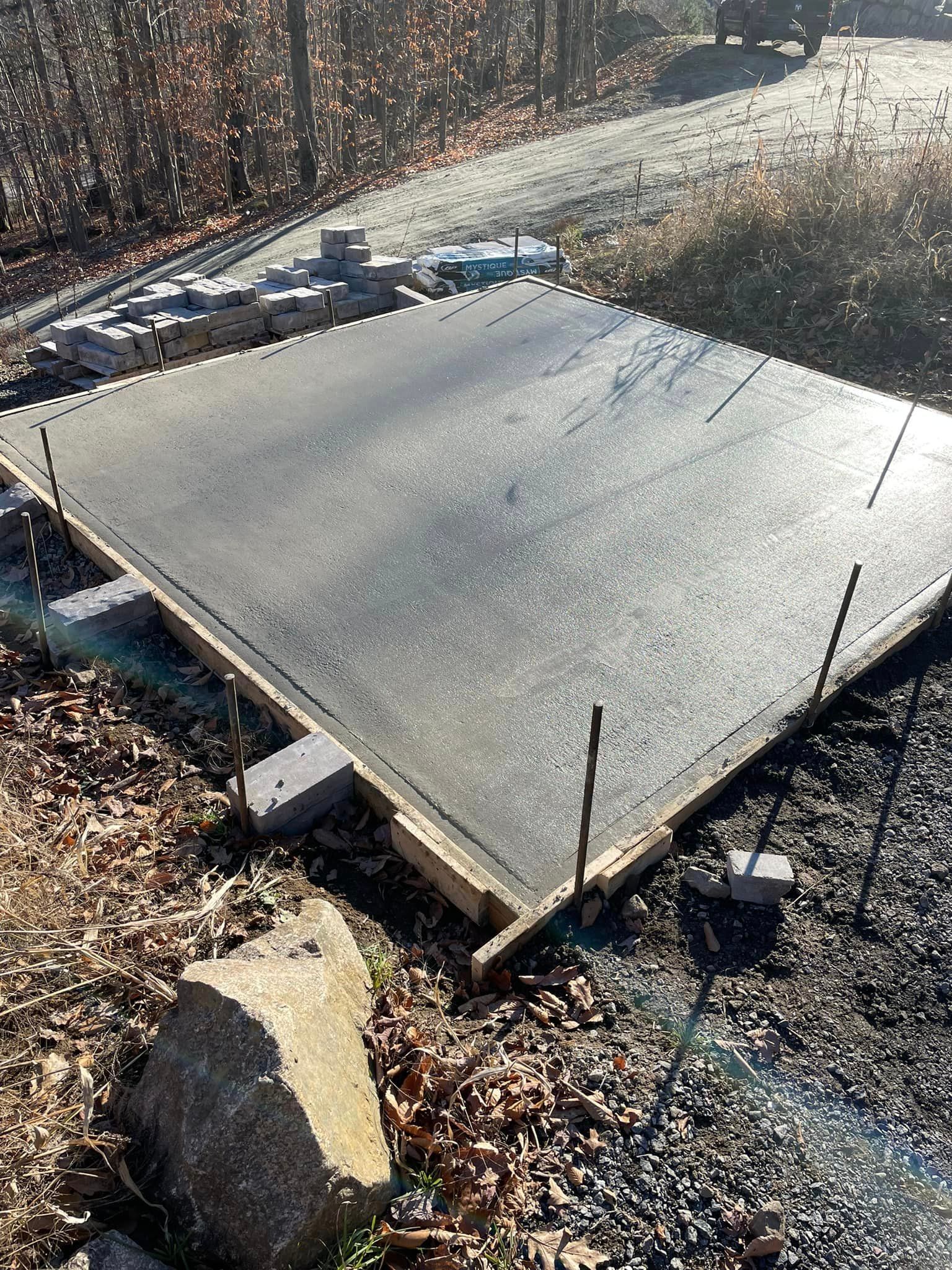 Dalle de béton fraîchement coulée dans une zone boisée, entourée d'une structure en bois, de briques et d'armatures.