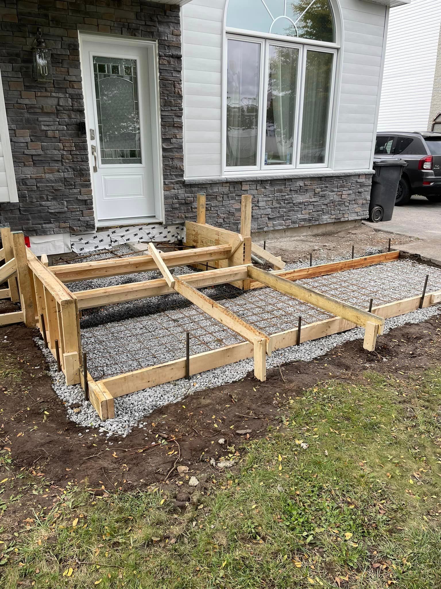 Coffrages en bois avec base de gravier, probablement destinés à une rampe en béton, devant une maison.