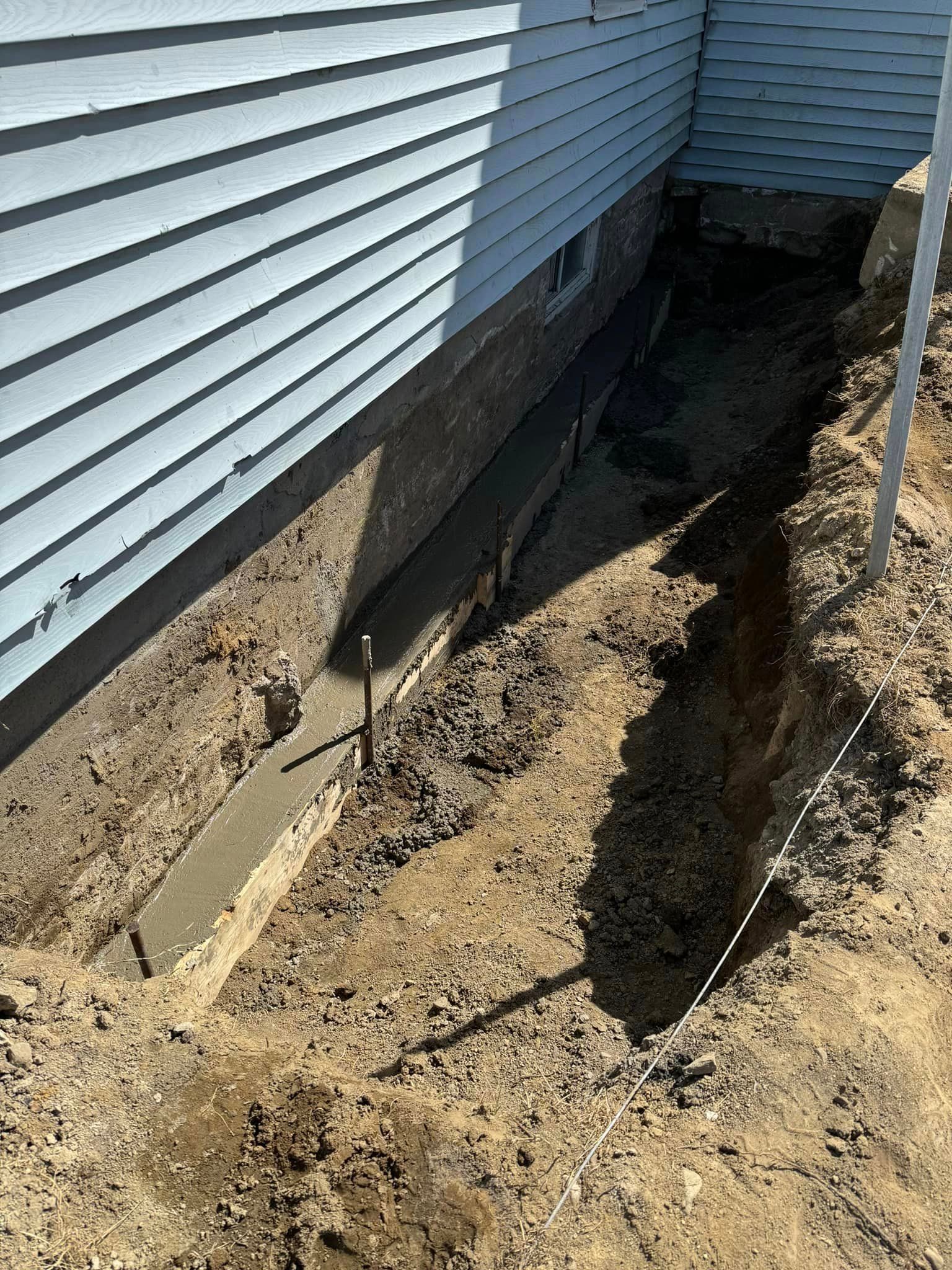 On coule des fondations en béton le long d'une maison bleu clair. La tranchée est entourée de terre.