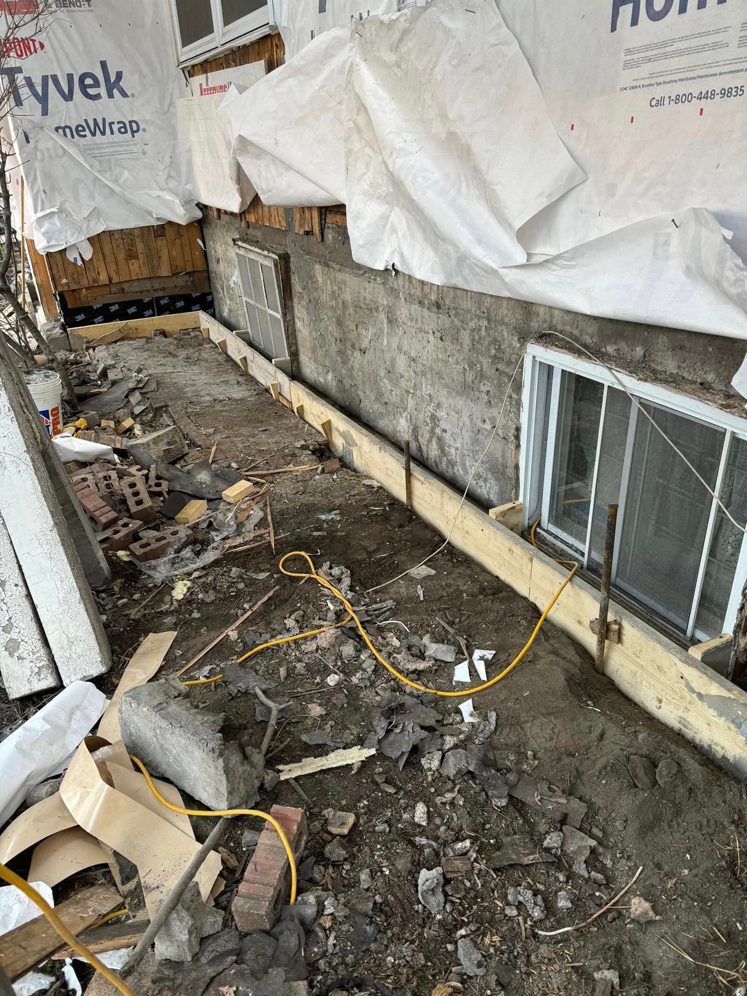 Chantier : fondations, fenêtres, bâche Tyvek, terre, débris et coffrages en bois.
