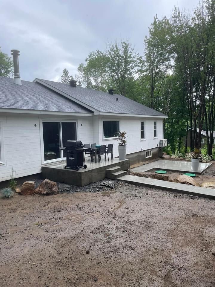 Maison blanche avec terrasse en béton, barbecue et mobilier de jardin. Arbres et ciel nuageux en arrière-plan.