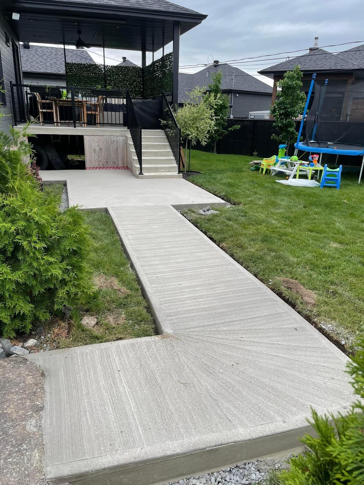 Allée en béton menant à une terrasse surélevée avec escalier, dans un jardin.