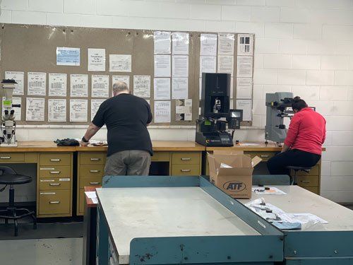 Two workers at a wall-mounted workbench in a workshop, facing blueprints and a machine.