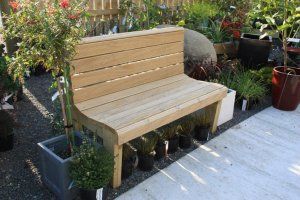 A light-colored wooden garden bench sits on a gravel patio surrounded by various potted plants and shrubs.