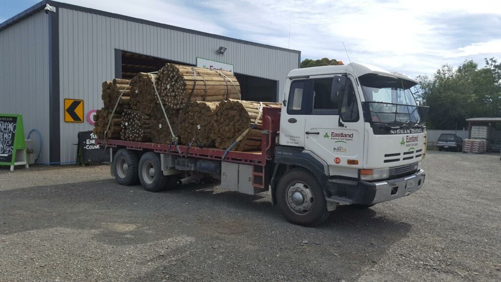 A white flatbed truck parked in front of a metal warehouse, loaded with large, cylindrical bundles of wooden poles.