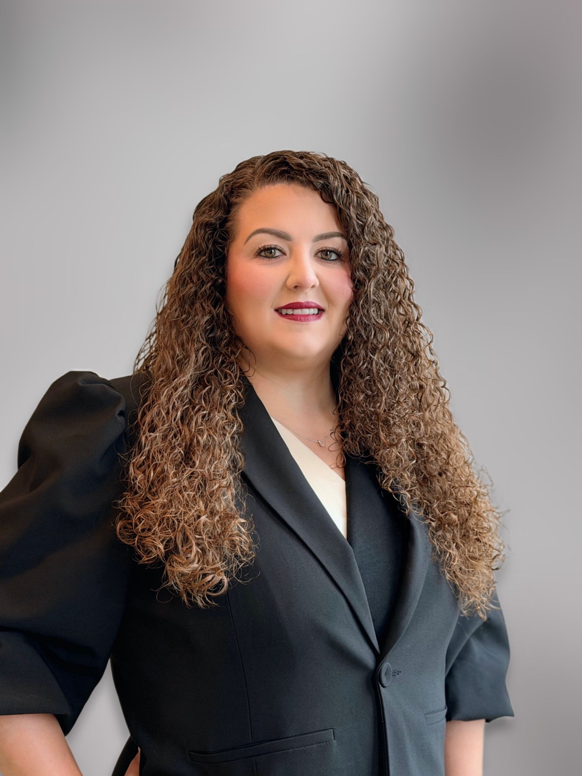 Vrouw met lang krullend haar in een bordeauxrode blouse en zwarte blazer tegen een donkergrijze achtergrond.