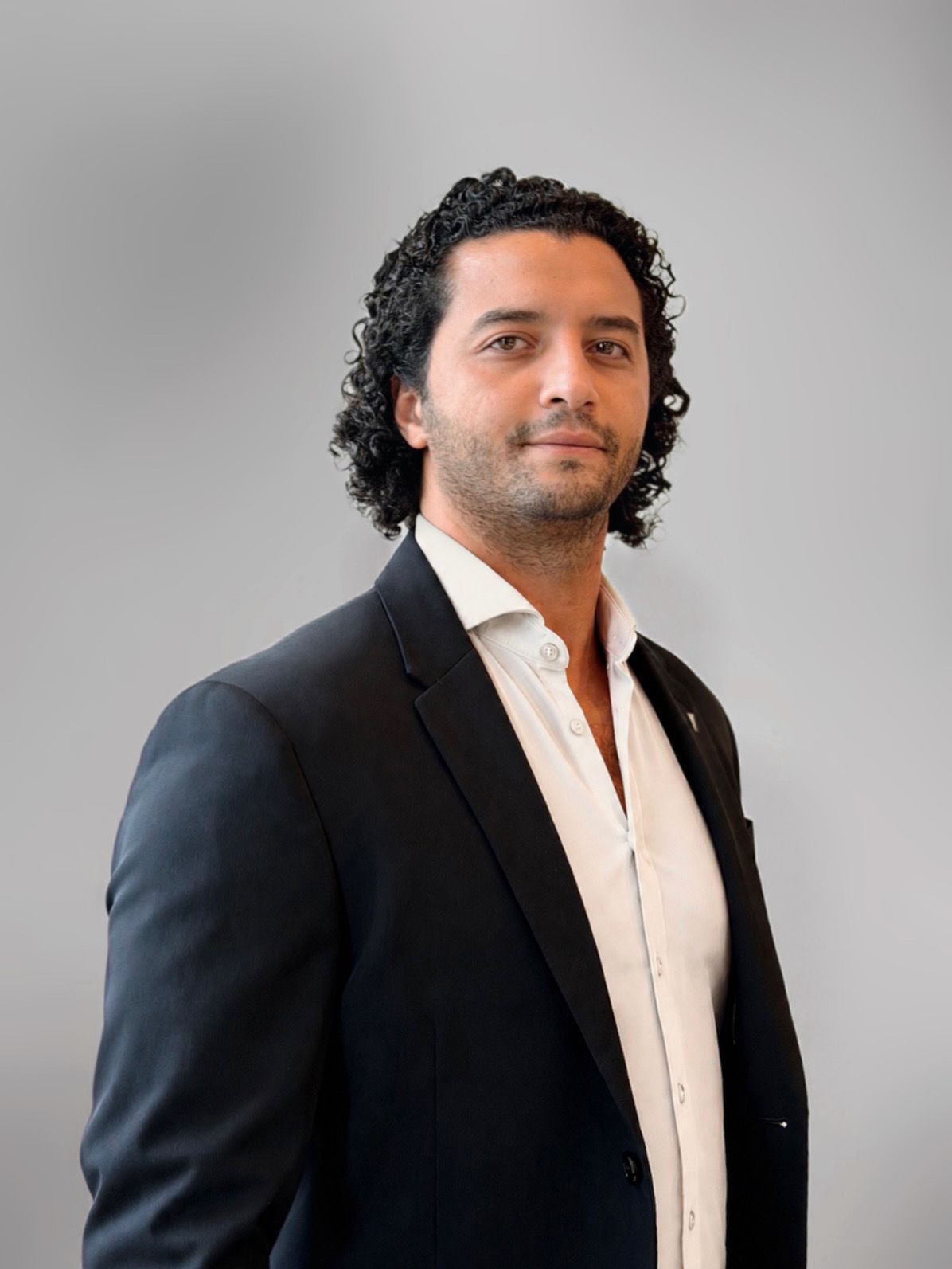 Een man in een grijze blazer en een zwart overhemd poseert tegen een effen grijze achtergrond.