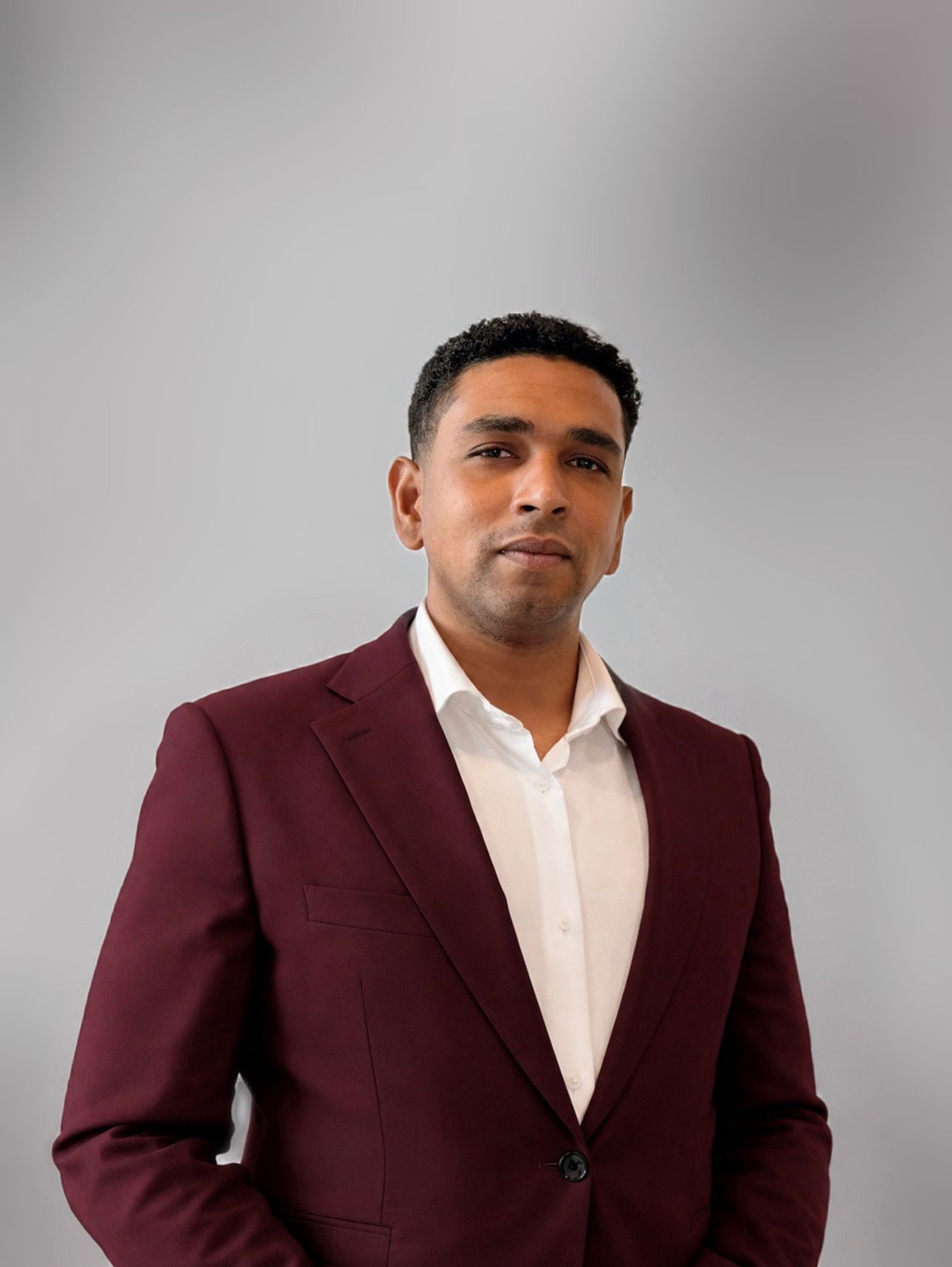 Een lachend persoon in een donkere blazer en een wit overhemd tegen een grijze achtergrond.