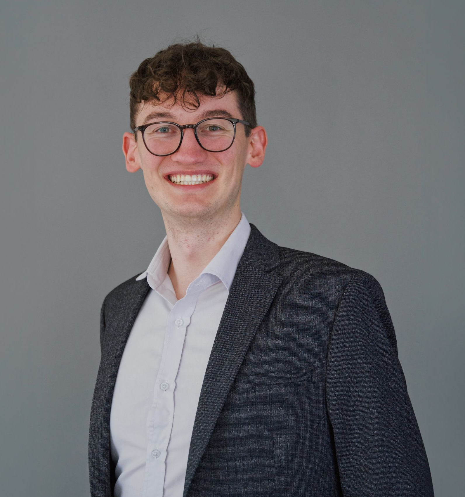 Lachende man met bril, gekleed in een wit overhemd en een donkere blazer, tegen een grijze achtergrond.