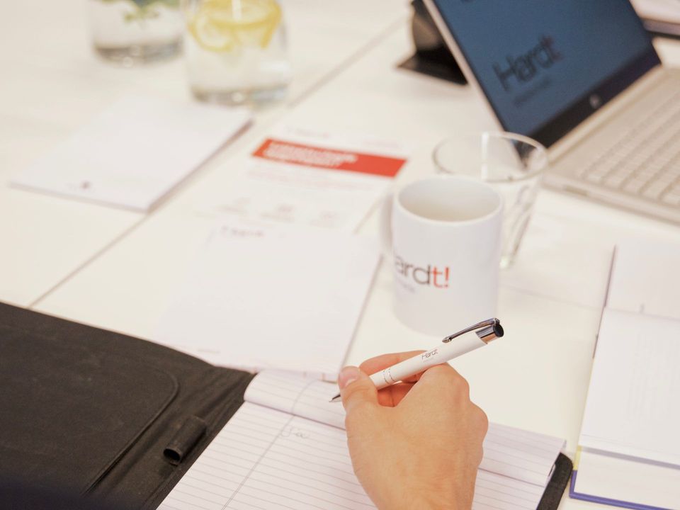 Een hand houdt een pen vast boven een notitieboekje op een bureau, met een witte mok en een laptop op de achtergrond.