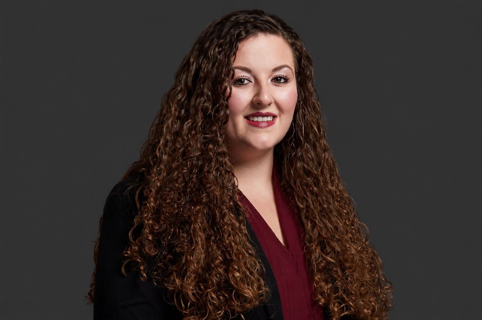 Vrouw met lang krullend haar in een bordeauxrode blouse en zwarte blazer tegen een donkergrijze achtergrond.