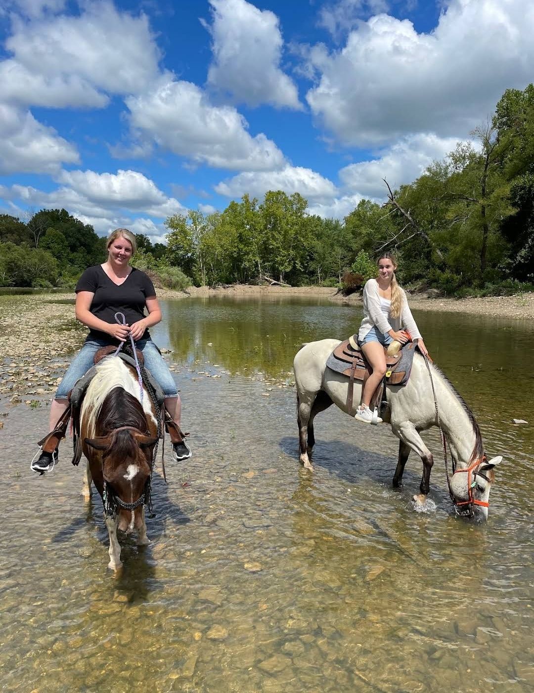 Two people riding horses through a shallow river under a bright, cloudy sky