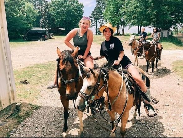 People horseback riding on a wooded trail, leading a line of horses through trees.